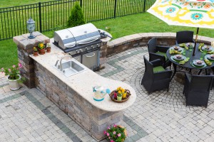 High angle view of a stylish outdoor kitchen gas barbecue and dining table set for entertaining guests with formal place settings and flowers on a paved patio